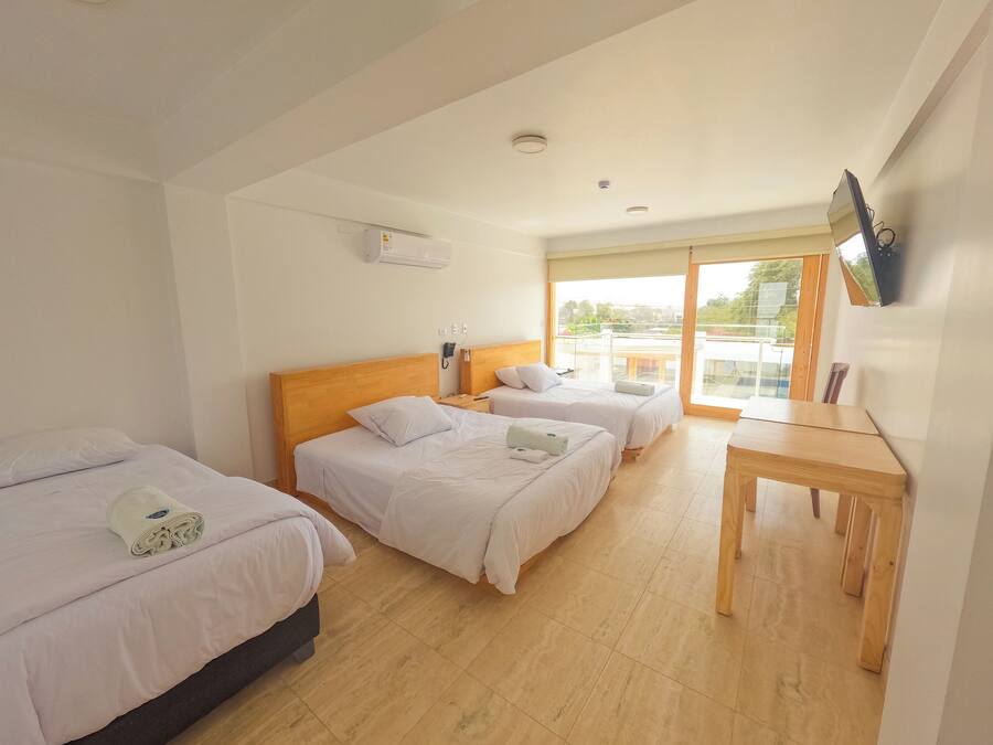 A hotel room with three beds, a wooden table, a TV mounted on the wall, and a window with a view of the outside.