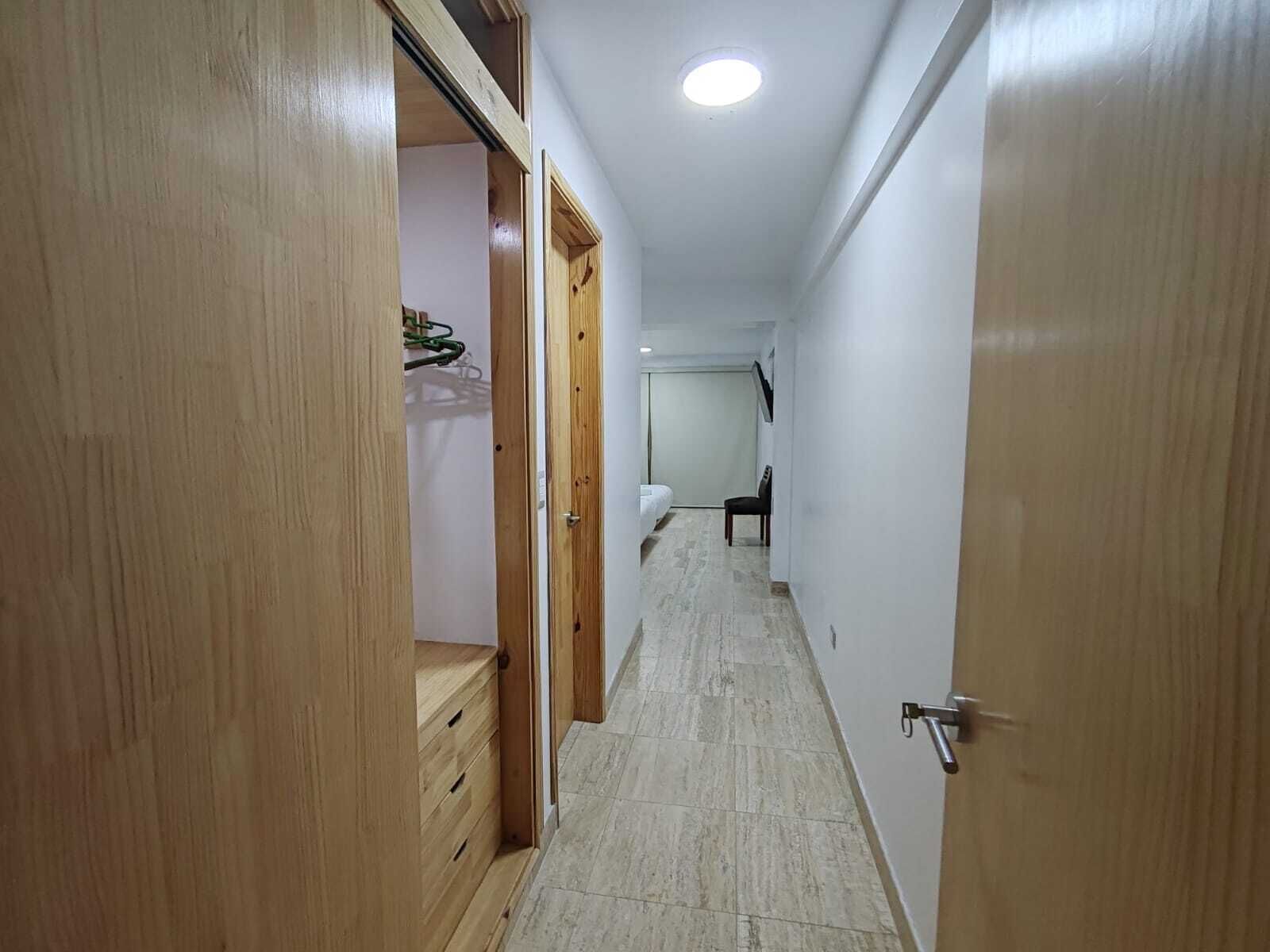 Un estrecho pasillo con puertas de madera a ambos lados, que conduce a una habitación con inodoro y una silla.