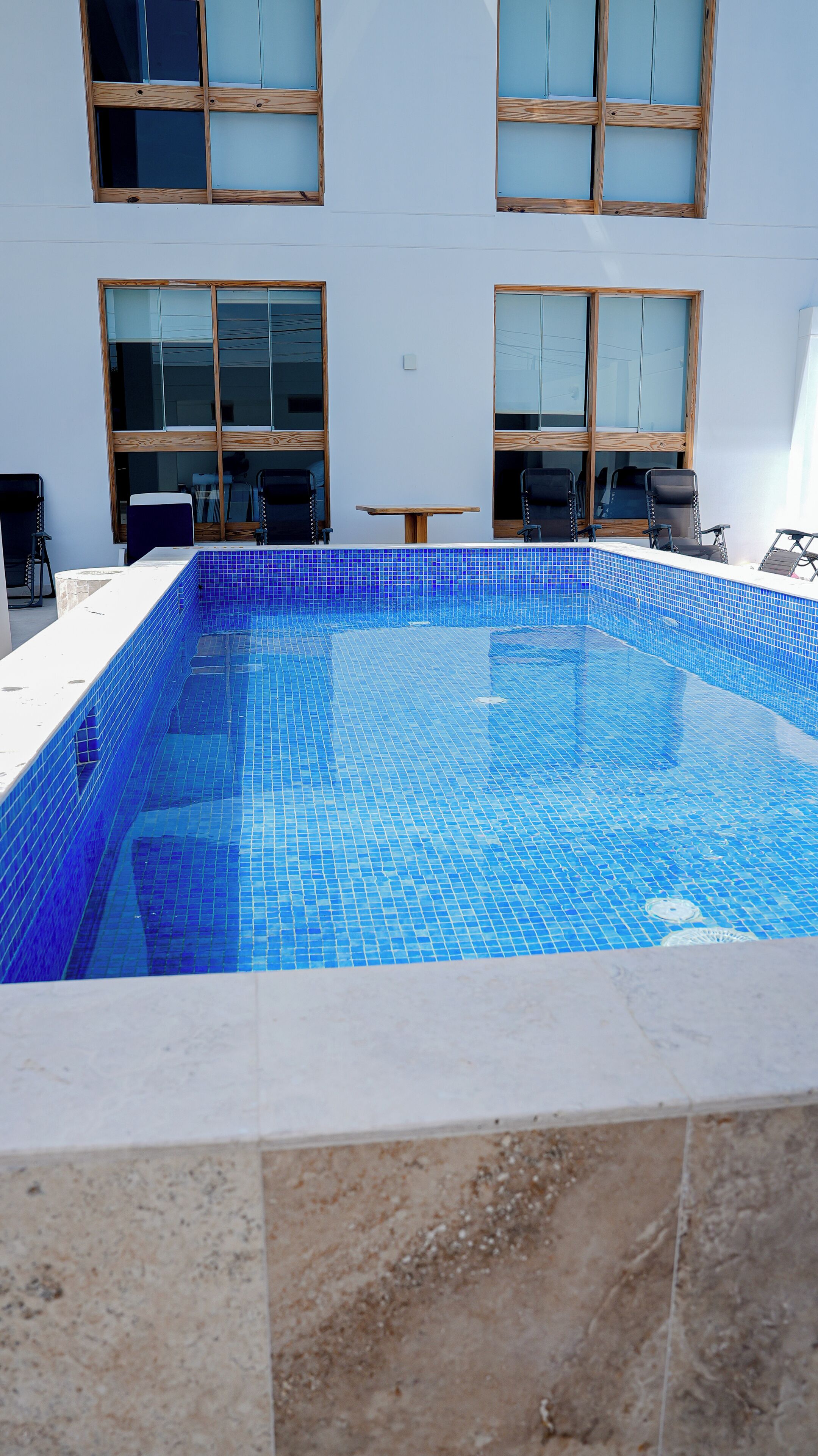 A rectangular swimming pool with blue mosaic tiles, surrounded by a white tiled deck.