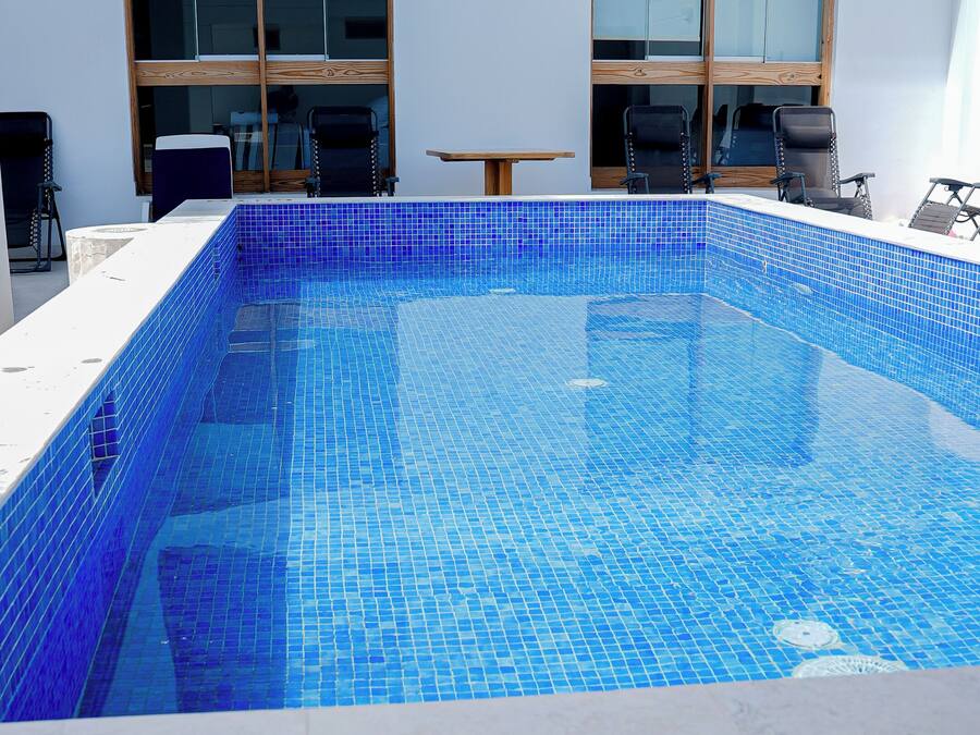A rectangular swimming pool with blue mosaic tiles, surrounded by a white tiled deck.