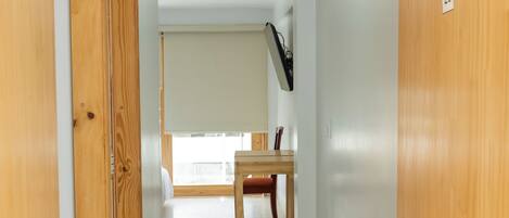 Un pasillo con una puerta de madera a la izquierda, una mesa y una silla de madera al fondo, y un techo blanco con una lámpara.