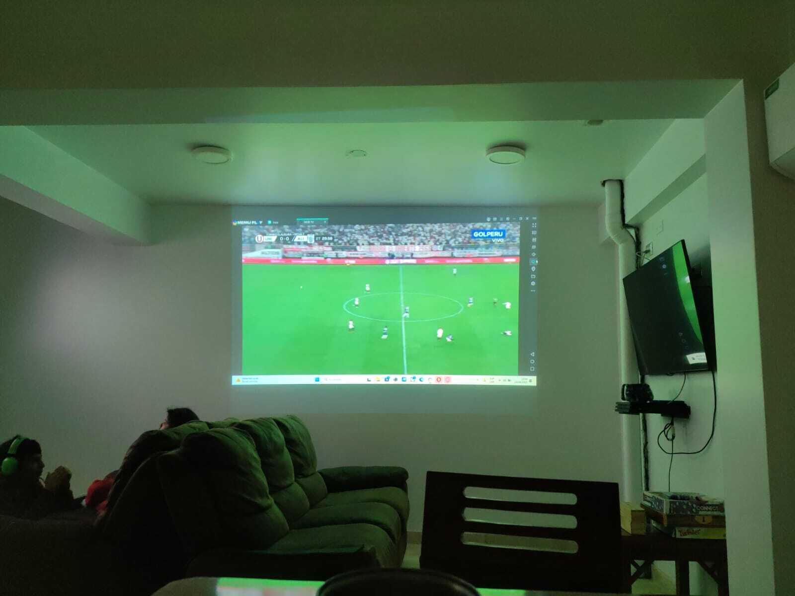 A room with a large screen displaying a soccer match, a couch, a chair, and a TV mounted on the wall.