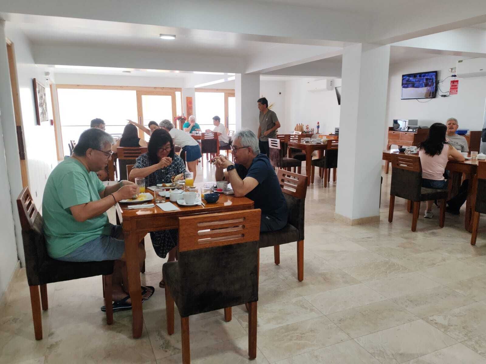 Un comedor con mesas y sillas de madera, un televisor montado en la pared y piso de baldosas.