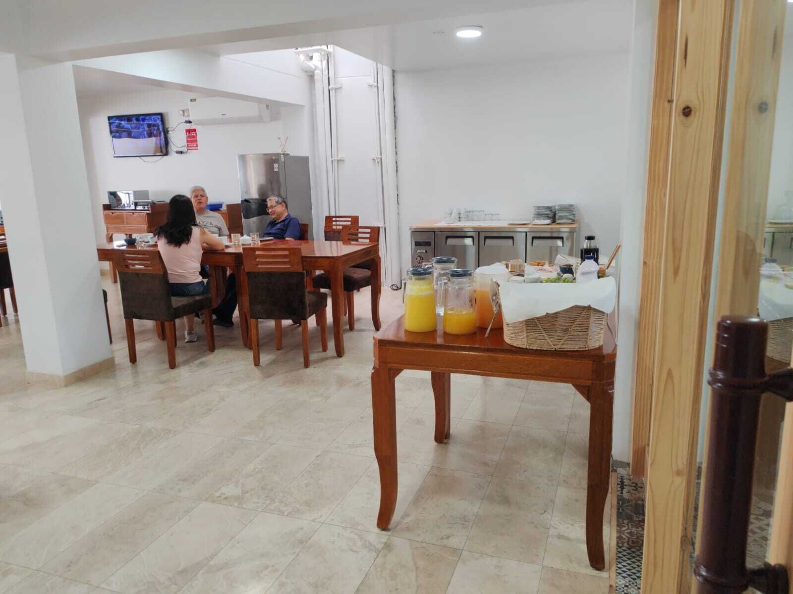 Un espace repas avec des tables et des chaises en bois, une cuisine équipée d’appareils électroménagers en acier inoxydable, et un buffet de petit-déjeuner proposant divers plats et boissons.