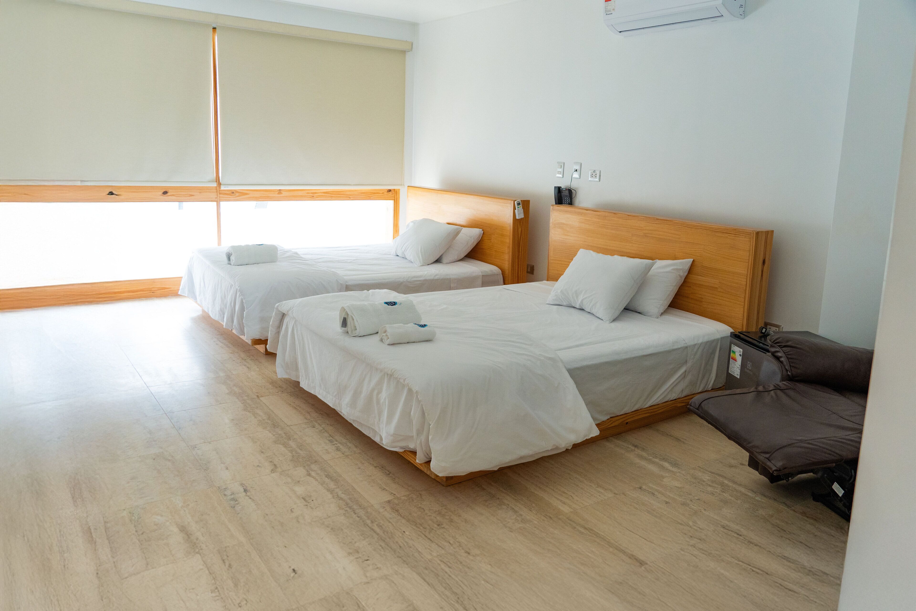 Habitación de hotel con dos camas individuales, un cabecero de madera, una mesita y una ventana con persianas.