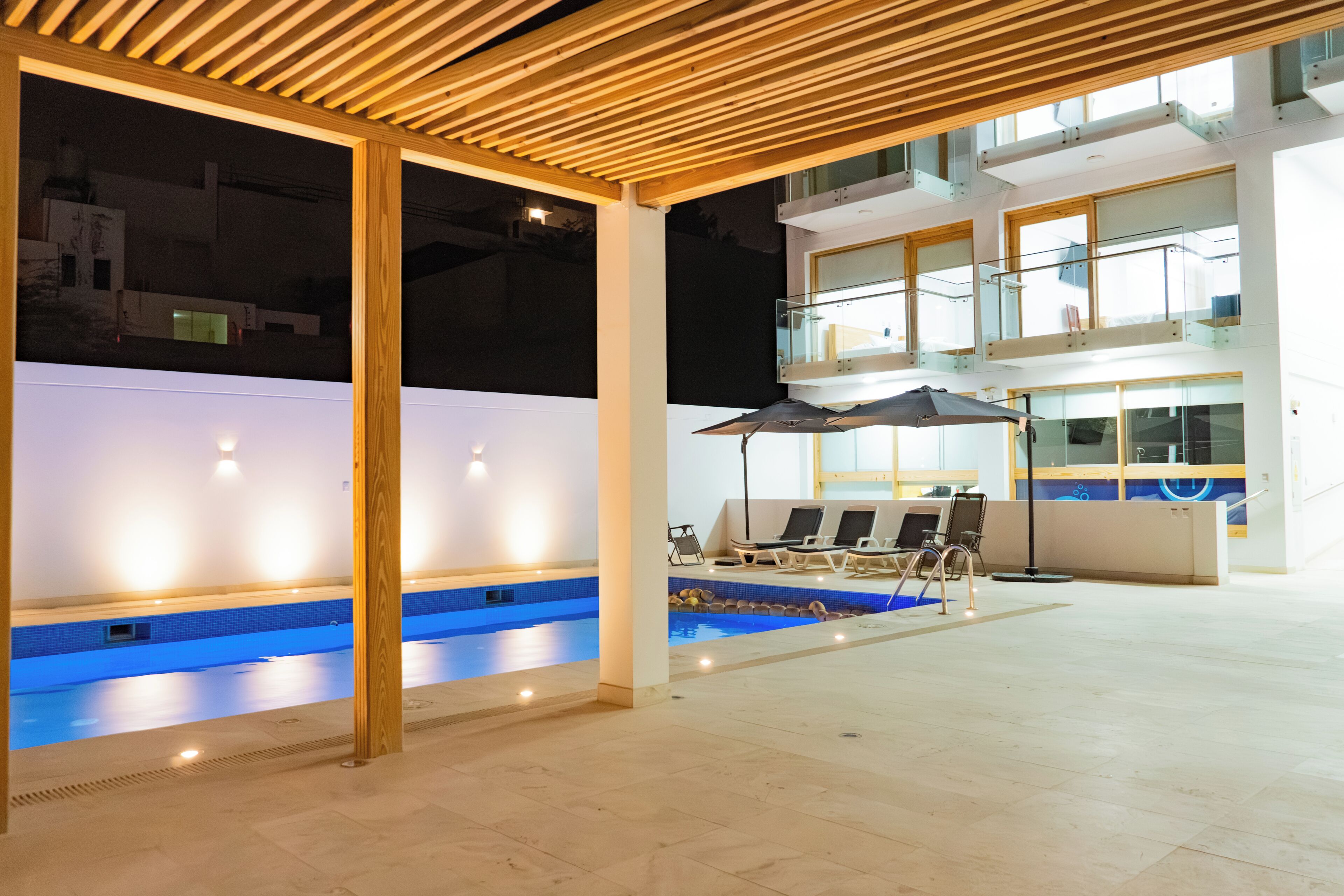 Ein moderner Innenbereich mit Schwimmbecken, verkleidet mit Holzbalken, Glasgeländern und einer Lounge-Ecke mit Sonnenschirmen.