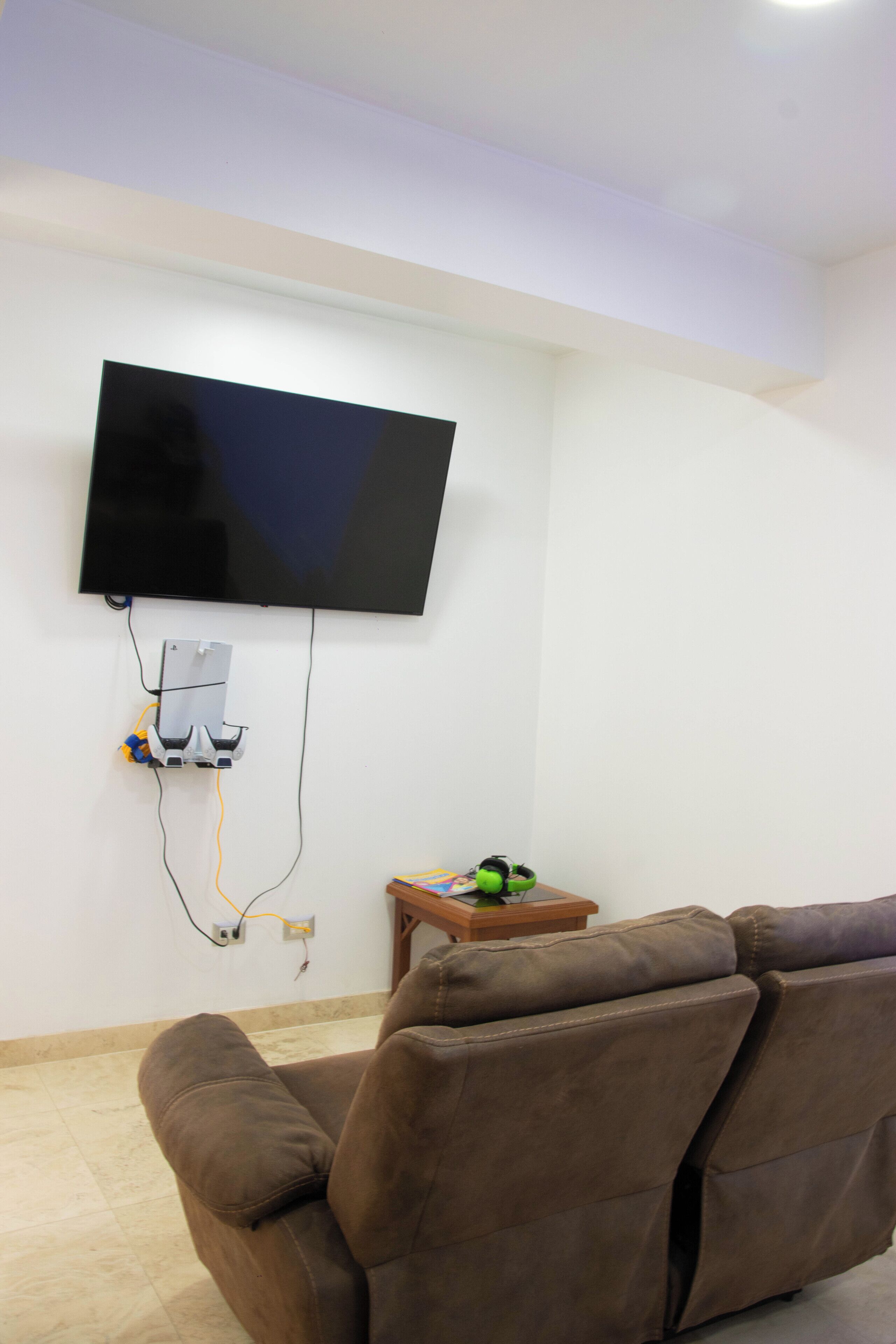 A living room with a flat-screen TV mounted on the wall, a brown sofa, and a small wooden side table with a green object on it.