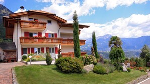 A two-story chalet with a wooden balcony, surrounded by lush greenery and mountains in the background.