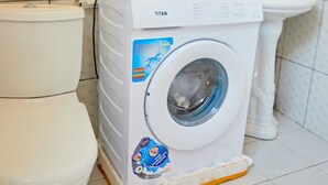 Lavadora blanca con una etiqueta, inodoro blanco y lavamanos blanco en un baño con paredes de azulejos.