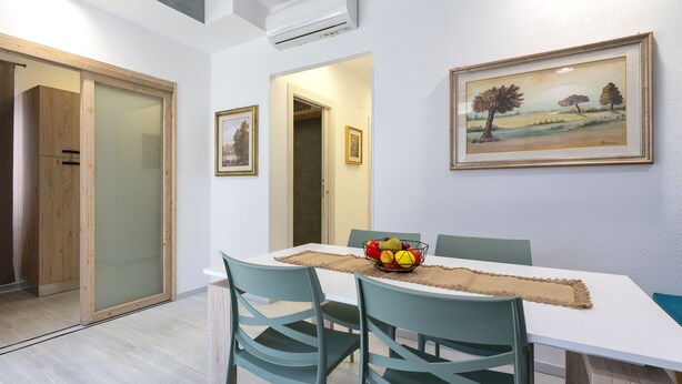 A dining area with a table, chairs, a fruit bowl, and framed artwork on the wall. - Domus Flavia by Interhome (Olbia)