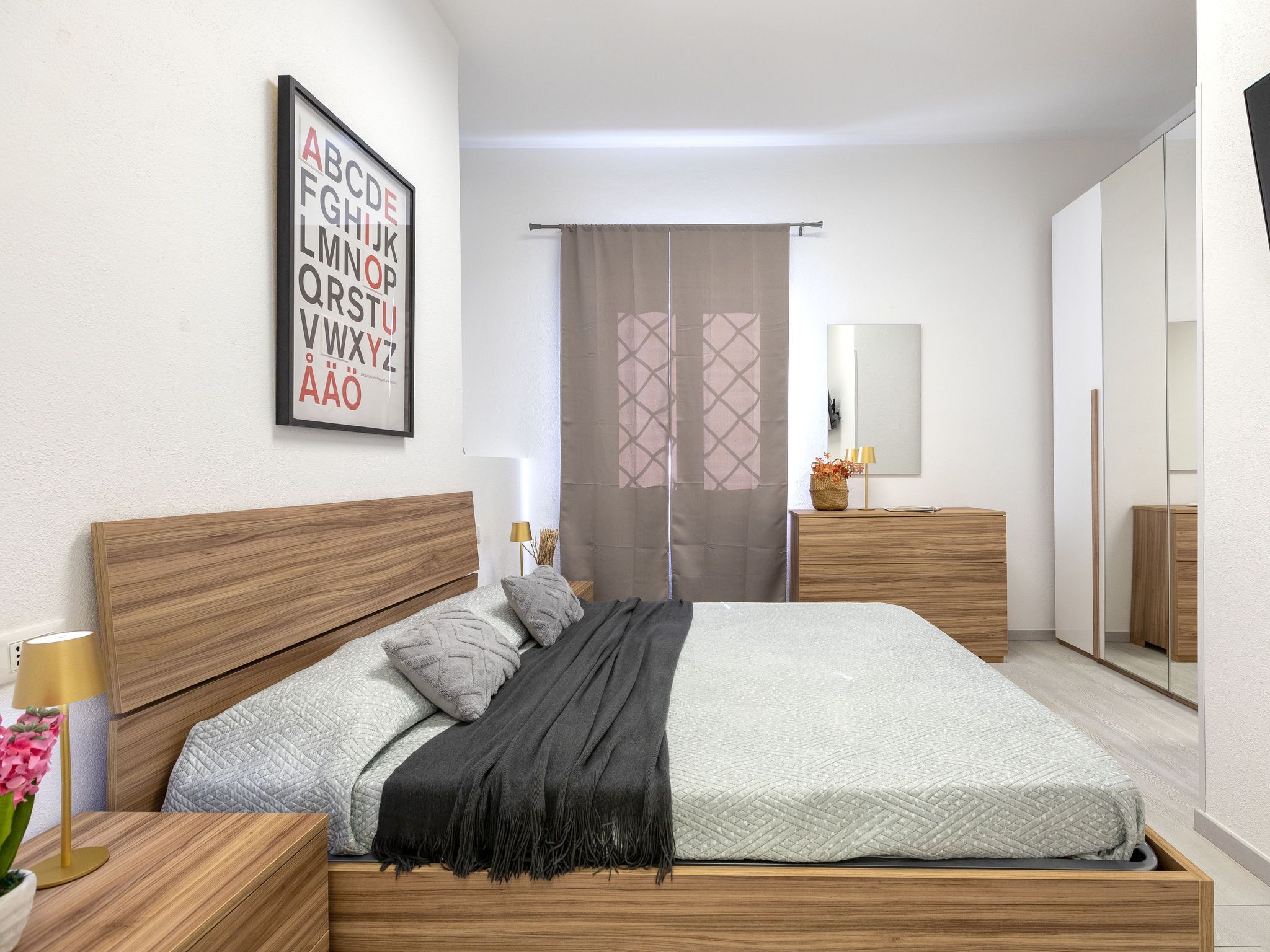 Ein modernes Schlafzimmer mit einem hölzernen Bett, einem Nachttisch mit Lampe, einem Spiegel und einem gerahmten Alphabet-Poster an der Wand.