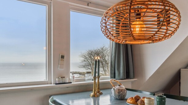 Een eetruimte met uitzicht op zee, een rieten hanglamp, een tafel met theepot, kaarsen en een bord gebak.