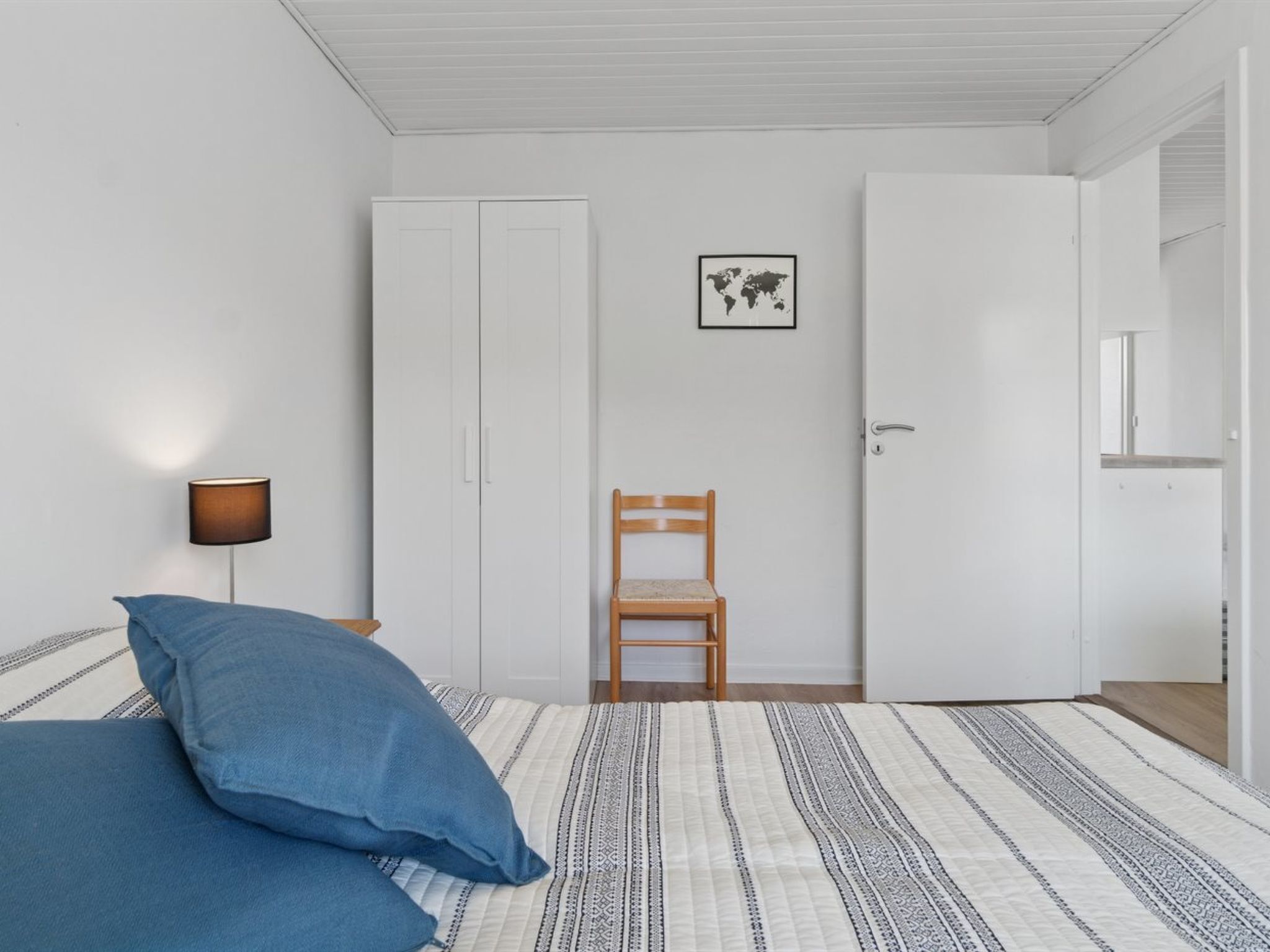 A bedroom with a bed, a chair, a wardrobe, a lamp, and a world map on the wall.