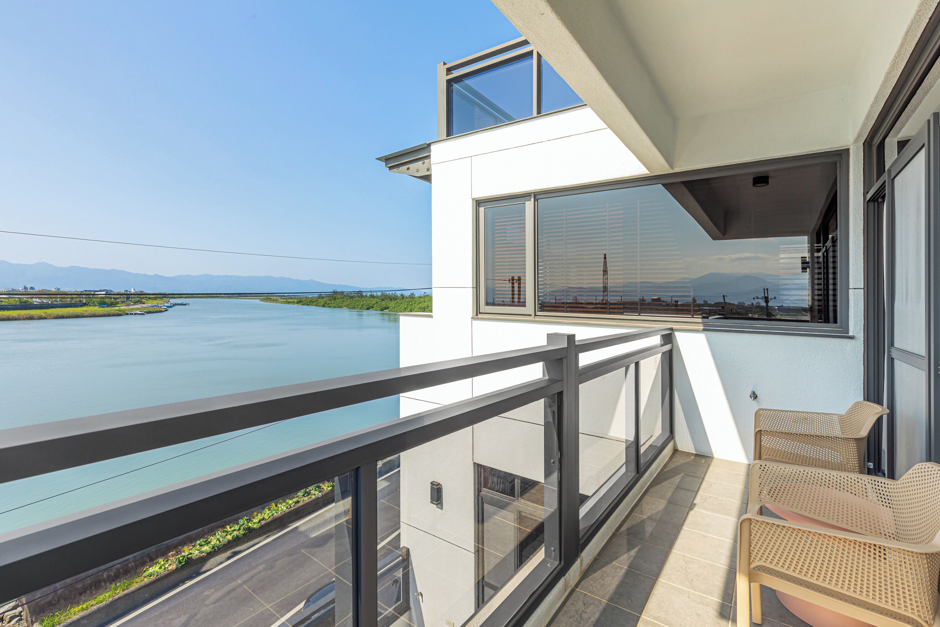 A balcony with a view of a river and mountains, wicker furniture, and a modern building facade.
