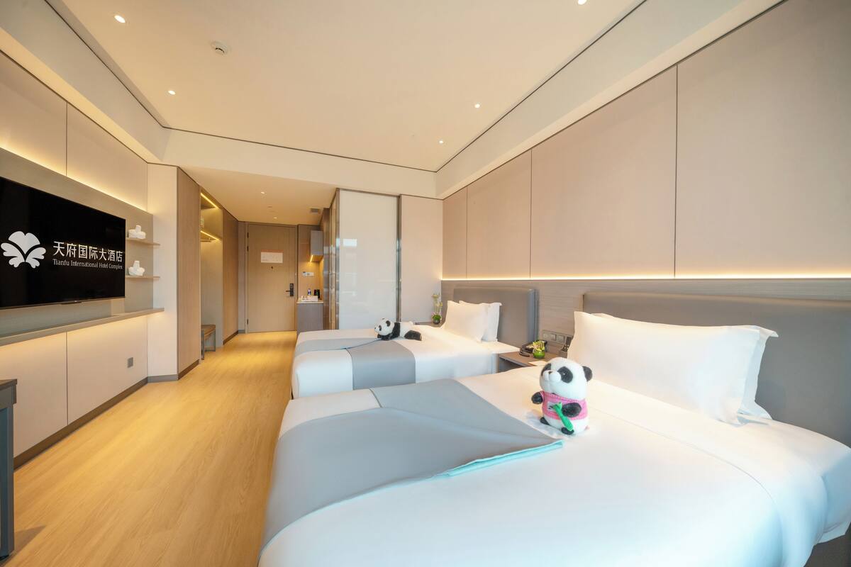 A modern hotel room with two beds, a flat-screen TV, and a panda plush toy on one bed.