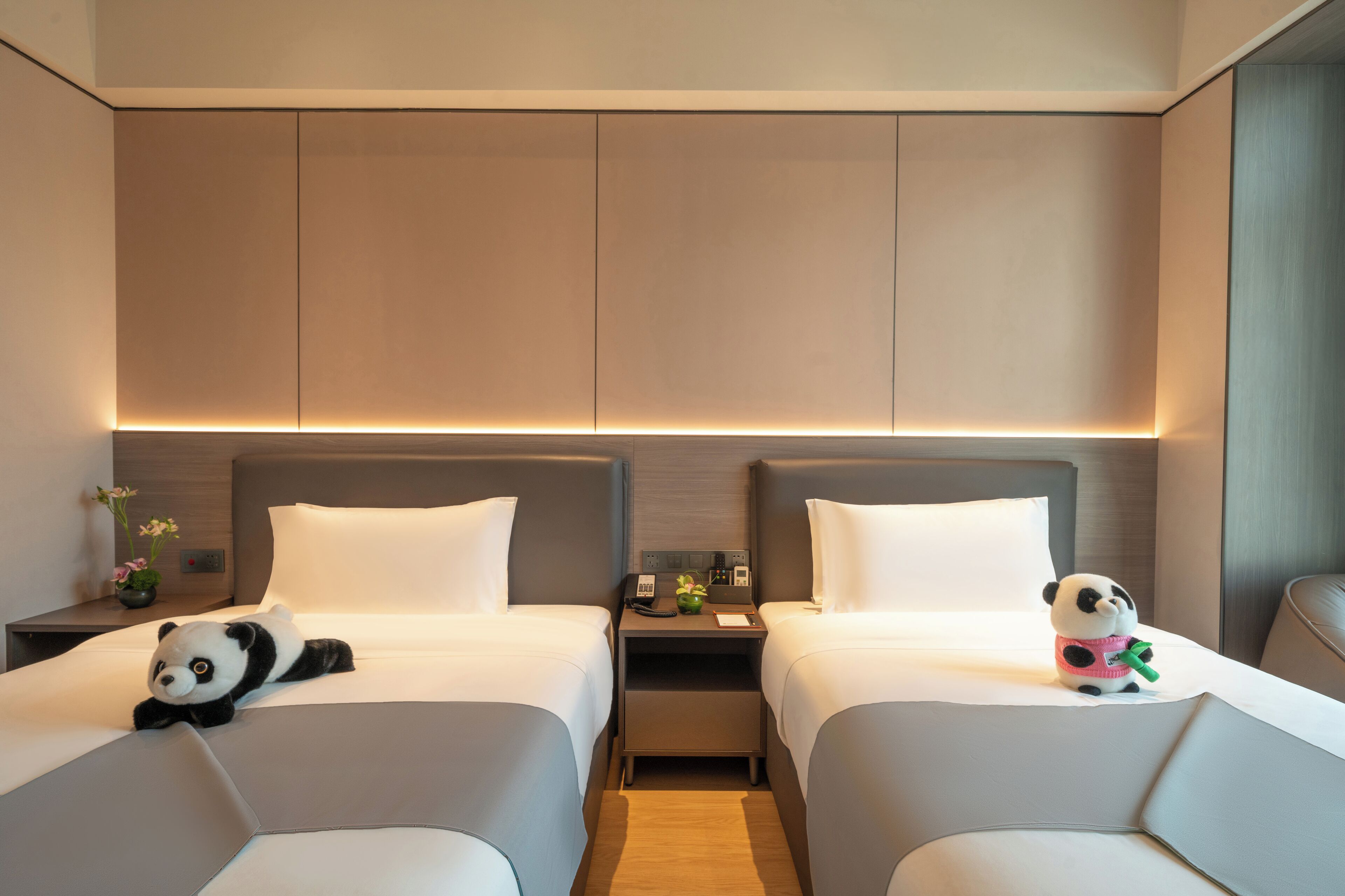 A hotel room with two beds, a nightstand with a lamp, a small table with a flower vase, and a panda plush toy on one bed.