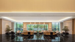 Un hall d’entrée spacieux au design moderne, comprenant un espace salon central avec des canapés et des fauteuils, de grandes fenêtres offrant une vue sur la verdure et un sol noir poli.