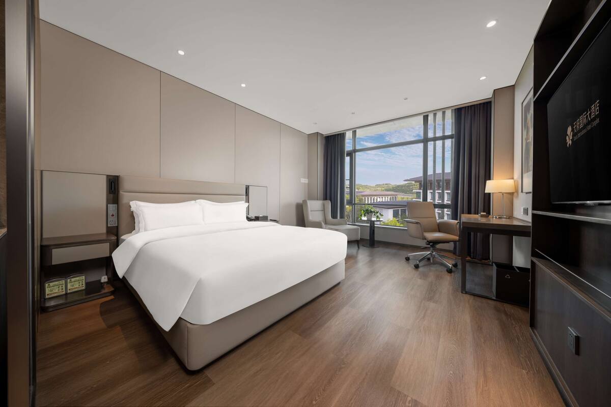 A modern hotel room with a large bed, a desk with a chair, a TV, and a large window offering a view of buildings.