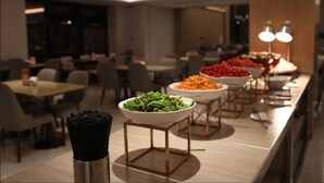 Un espace repas moderne avec plusieurs tables et chaises, un buffet proposant divers plats et un vase décoratif contenant des objets noirs.