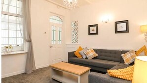A living room with a sofa, a coffee table, a lamp, and a window with curtains. - Kitty Cottage - 3 Bedroom - Warm and Welcoming - Sleeps 5 - Blackpool (Blackpool)