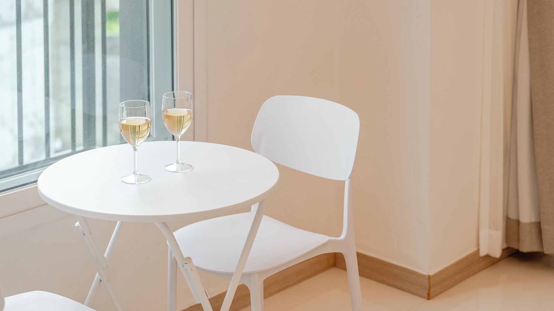 Une table blanche avec deux verres de vin blanc, une chaise blanche et une fenêtre avec des stores.