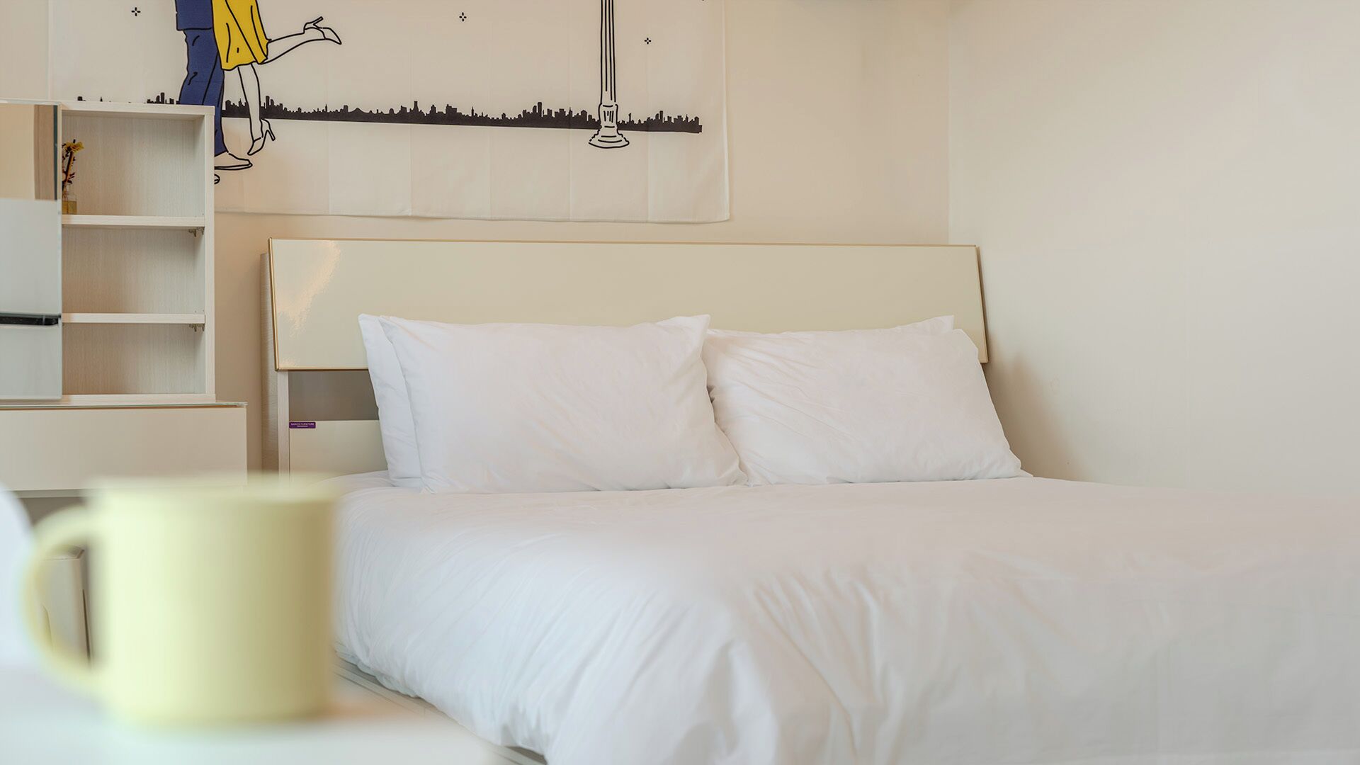 A bed with white bedding, a nightstand, a shelf with a vase, and a wall with a cityscape mural.