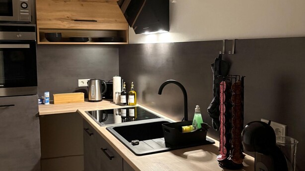 Une cuisine moderne équipée d’un four, d’un micro-ondes et d’un évier encastrés. On trouve un meuble en bois au-dessus de l’évier et une hotte noire.