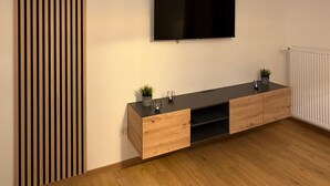 Un salon moderne avec un plancher en bois, un téléviseur noir fixé au mur et un meuble en bois contenant de la verrerie et une plante.
