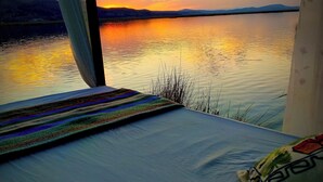 Ein Bett mit Blick auf einen Sonnenuntergang über dem Wasser, ein Fenster mit Vorhang und ein Kissen mit Muster.