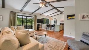 A spacious living room with a sofa, a coffee table, and a dining area with chairs. There is a kitchen area with a TV and a chandelier.