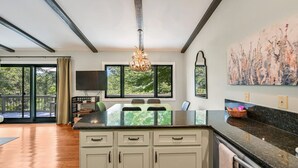 A modern kitchen with a large island, dark countertops, and white cabinetry. There are two windows with a view of greenery outside, and a chandelier hanging above the island.