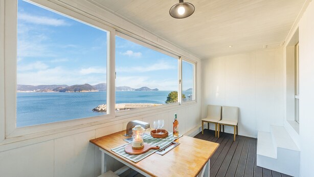 A room with a wooden table, a bottle, and a glass, with a view of the sea and hills. - Geoje Kkachi and Santorini Pension (Geoje)