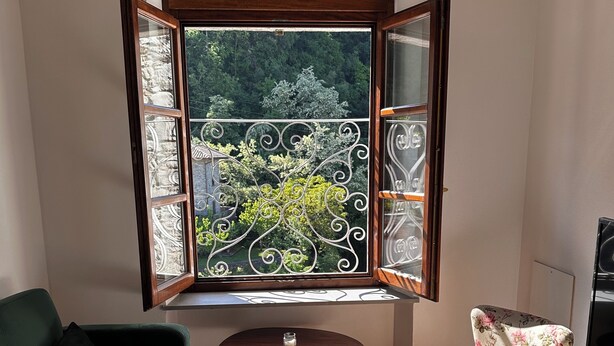 A small room with a green sofa, a floral chair, a small table, and a cabinet. A window with decorative iron bars offers a view of greenery outside. - Il suono del fiume (Bagnone)