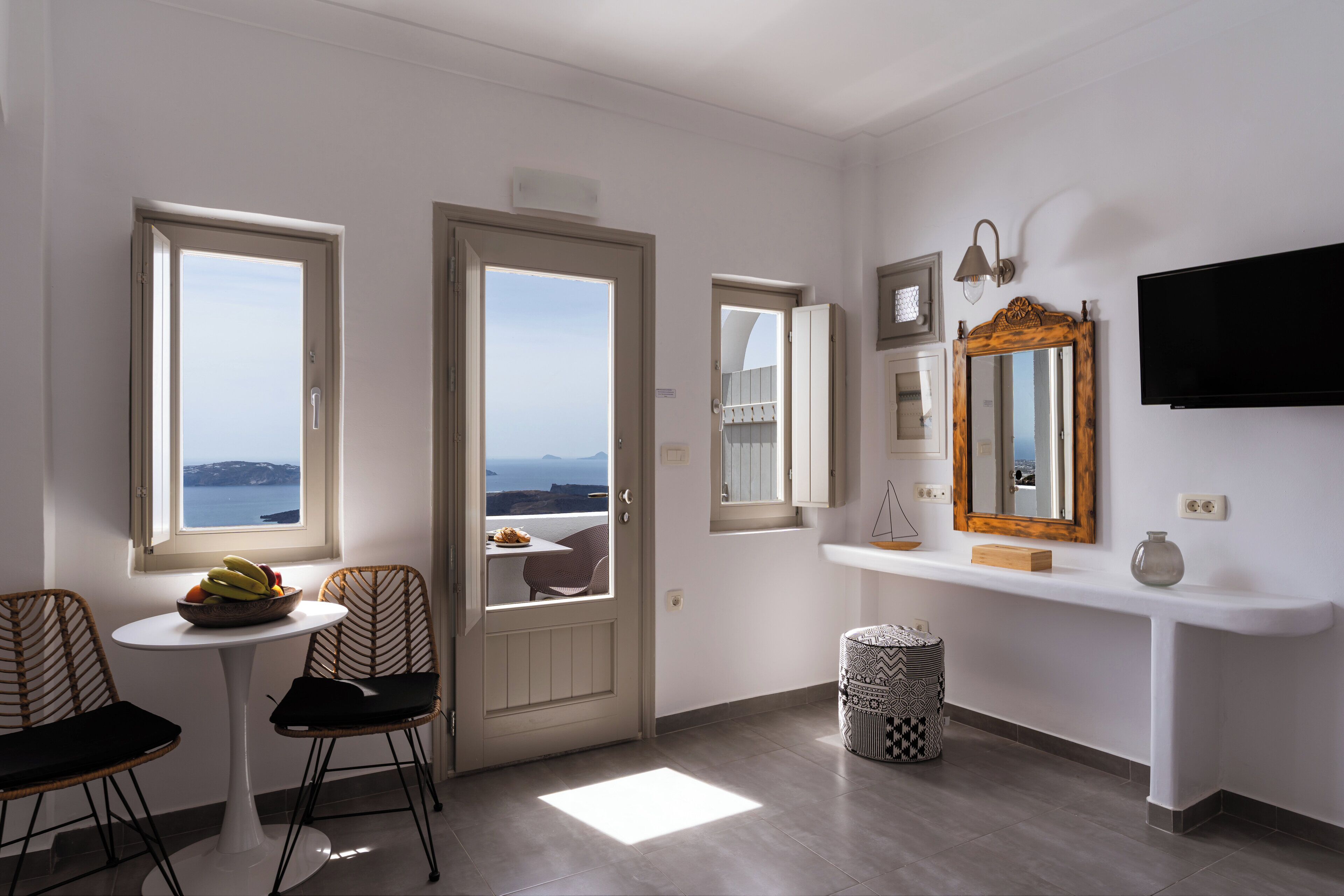 A room with a view of the sea, a dining area with chairs and a table, a white console table with a mirror and a TV, and a patterned waste basket.