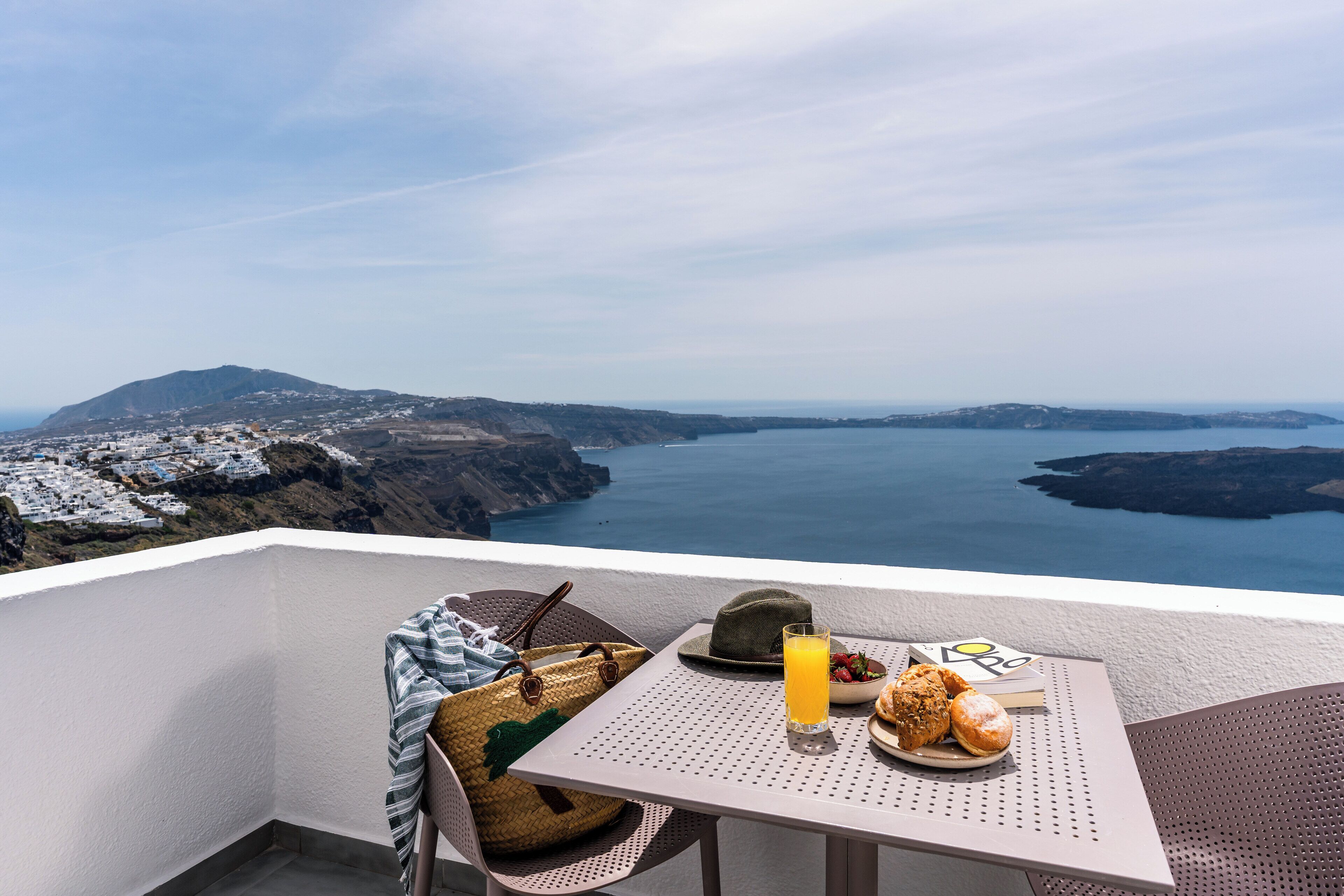 A balcony with a table set for breakfast, overlooking a coastal landscape with white buildings and a view of the sea.