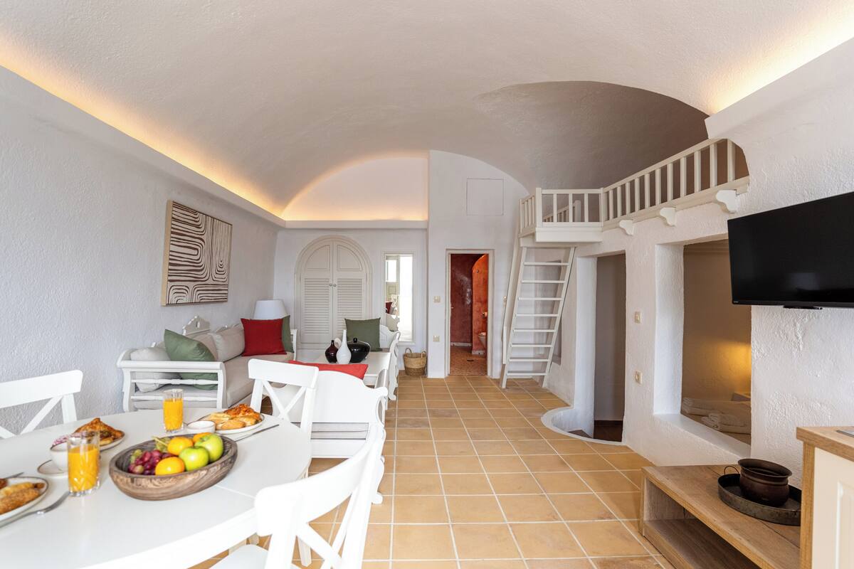 Amplia sala de estar con una mesa de comedor lista para el desayuno, un televisor montado en la pared y una escalera que lleva a un nivel superior.