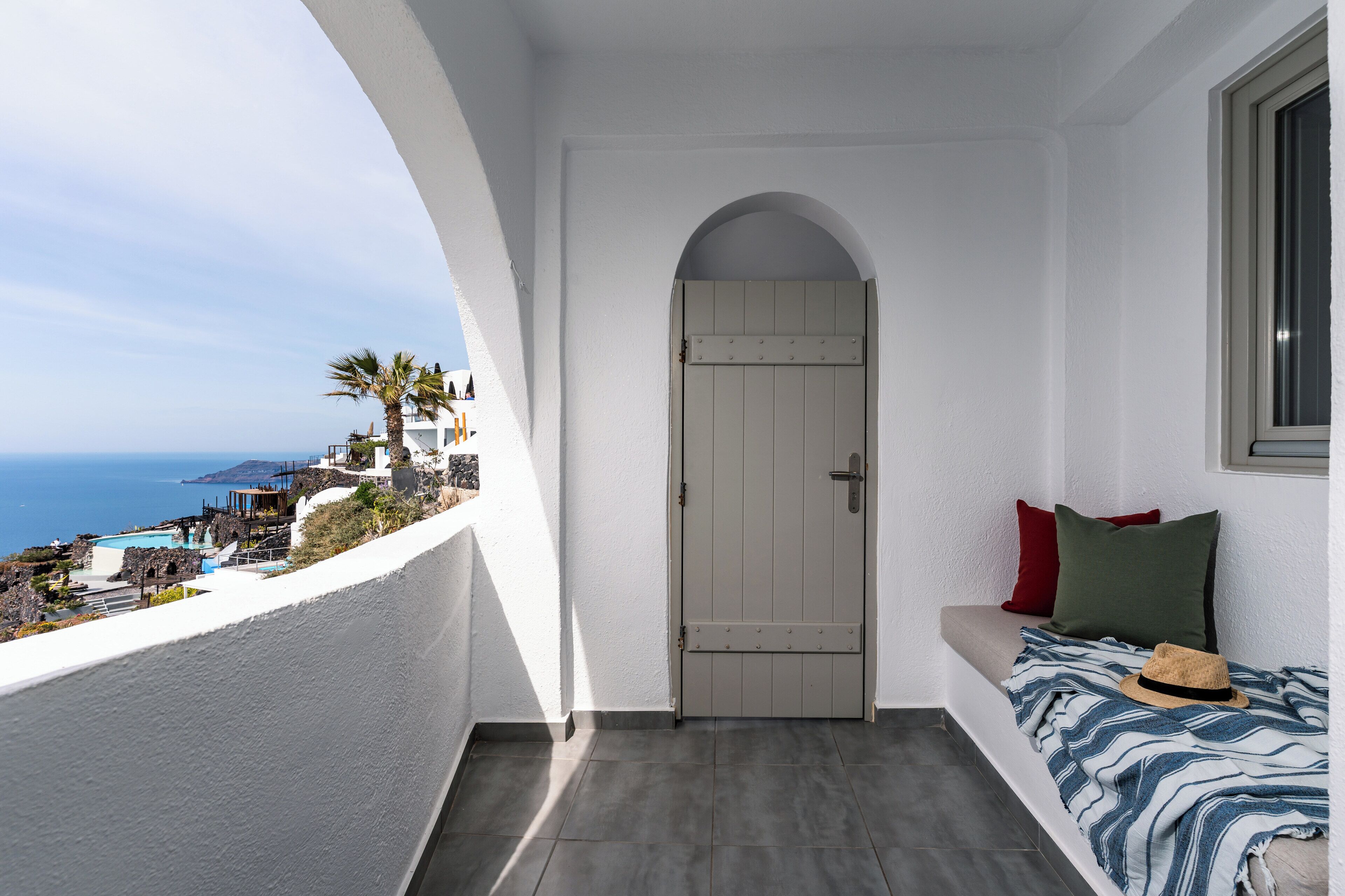 A balcony with a view of the sea, a white door, and a bed with a striped blanket and a hat.