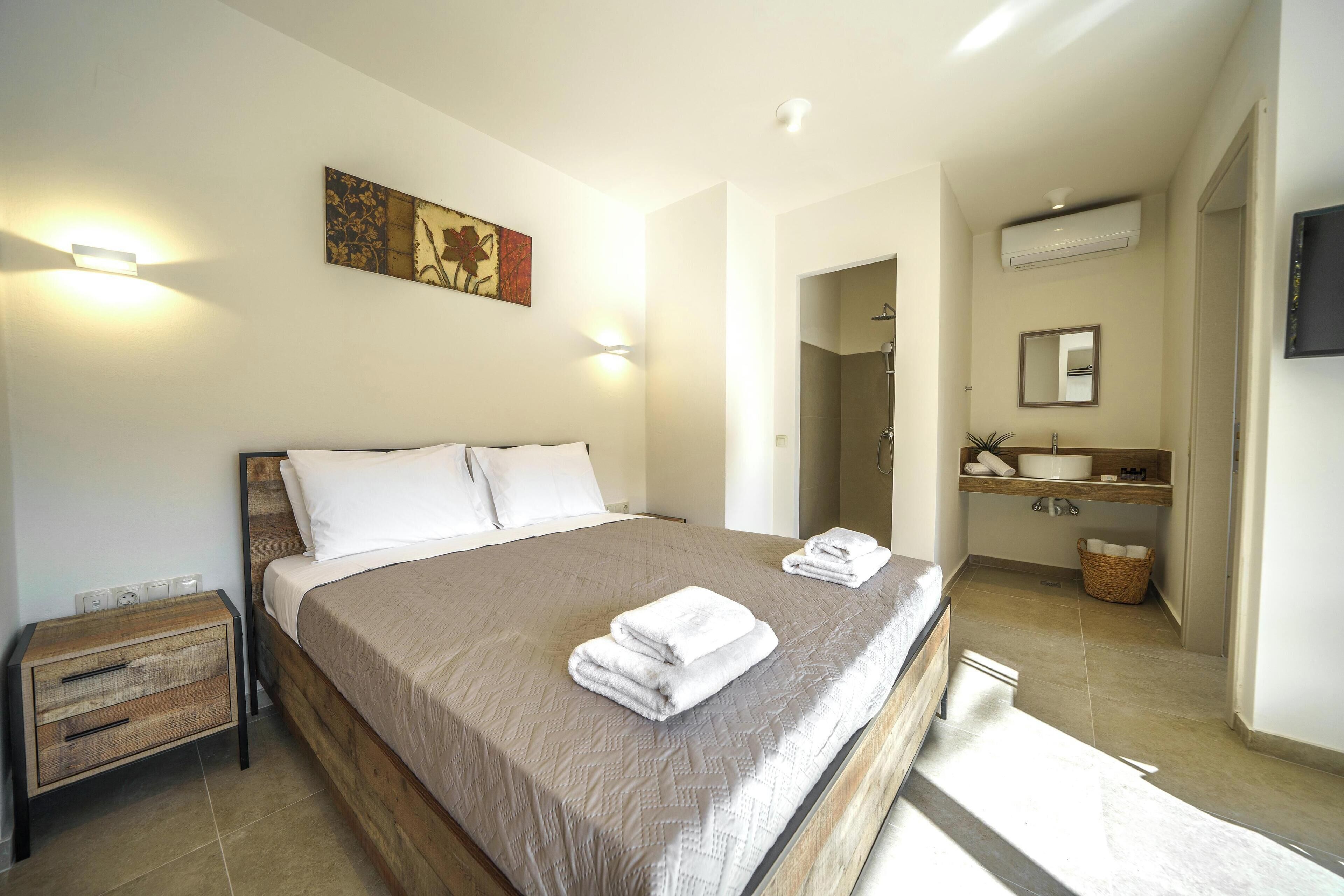 Un dormitorio con una cama grande, mesitas de noche, una obra de arte en la pared, un baño con lavabo y espejo, y un equipo de aire acondicionado.