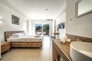 Un dormitorio moderno con una cama de madera, un ventanal grande, un baño con lavamanos y espejo, y un televisor en la pared.