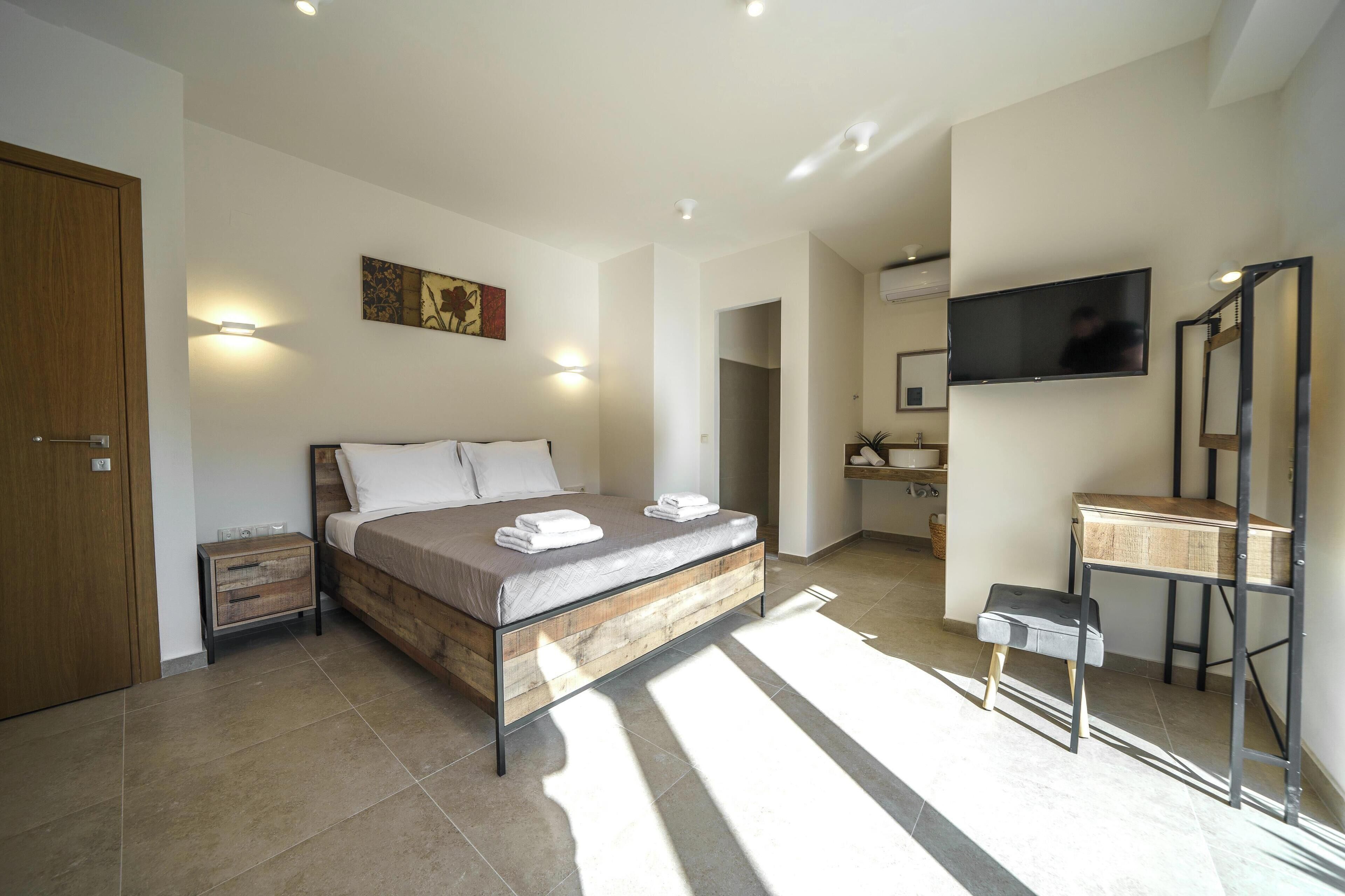A modern bedroom with a bed, bedside tables, a TV, and a bathroom visible in the background.