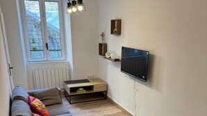 A modern living room with a grey sofa, a small ottoman, a wooden coffee table, a TV mounted on the wall, and a window with a view of the outside.