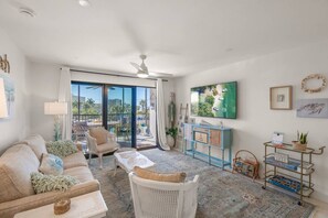 A living room with a sofa, armchair, coffee table, and a TV on a wall. There is a sliding glass door leading to an outdoor area. - Harbor Towers | Stunning Siesta Key Water Views! (Siesta Key)