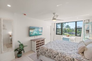 A bedroom with a large bed, a ceiling fan, a TV mounted on the wall, a window with a view of palm trees and a road, and a small desk with a chair. - Beautifully Renovated Ocean View Condo at Harbor Towers | Walk to Beach | Heated Pools & Tennis (Siesta Key)
