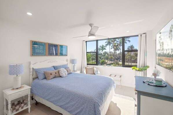 A bedroom with a bed, bedside tables, a ceiling fan, and a large window with a view of palm trees.
