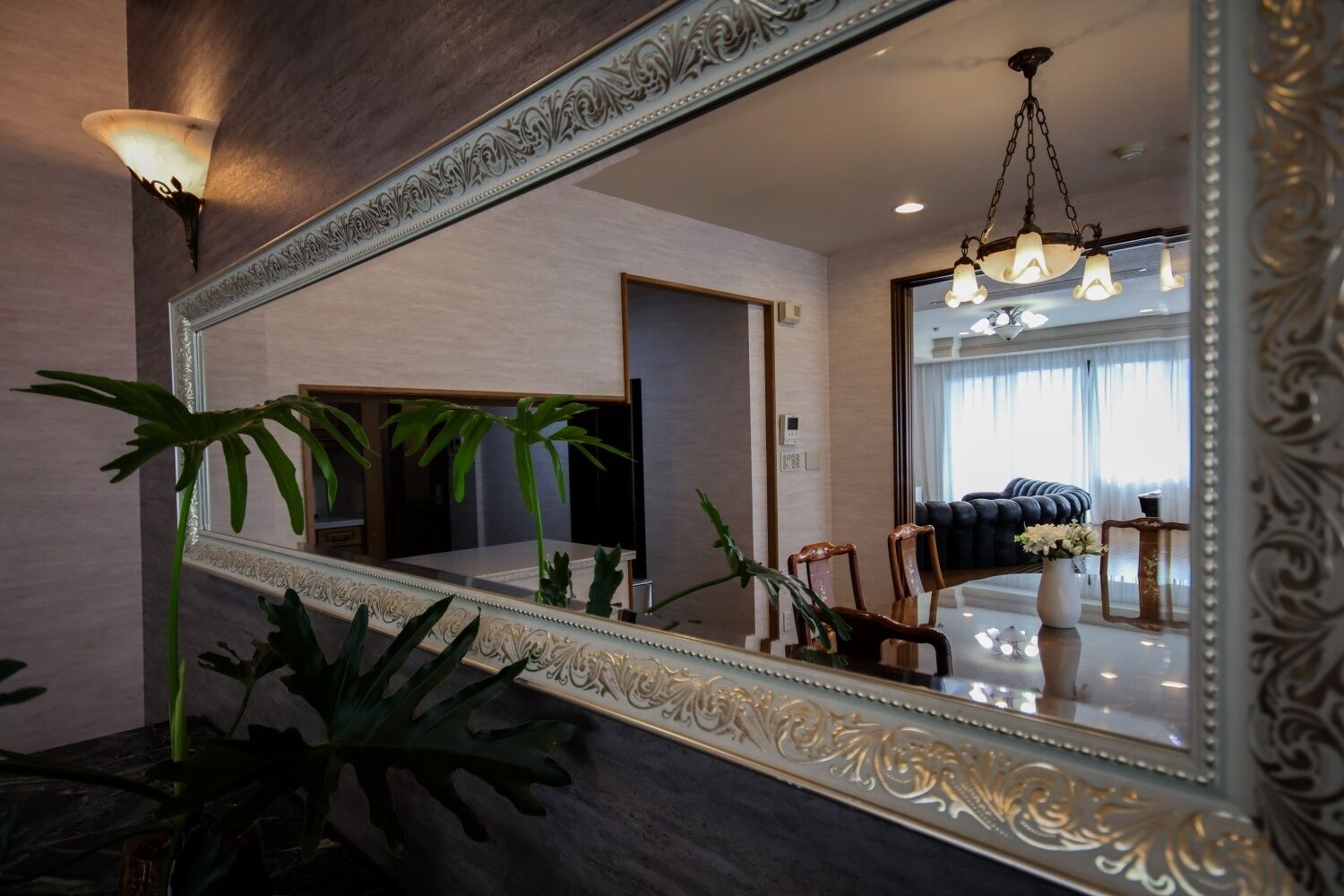 Un grand miroir décoratif reflète un salon moderne avec un canapé foncé, un lustre et un coin repas.