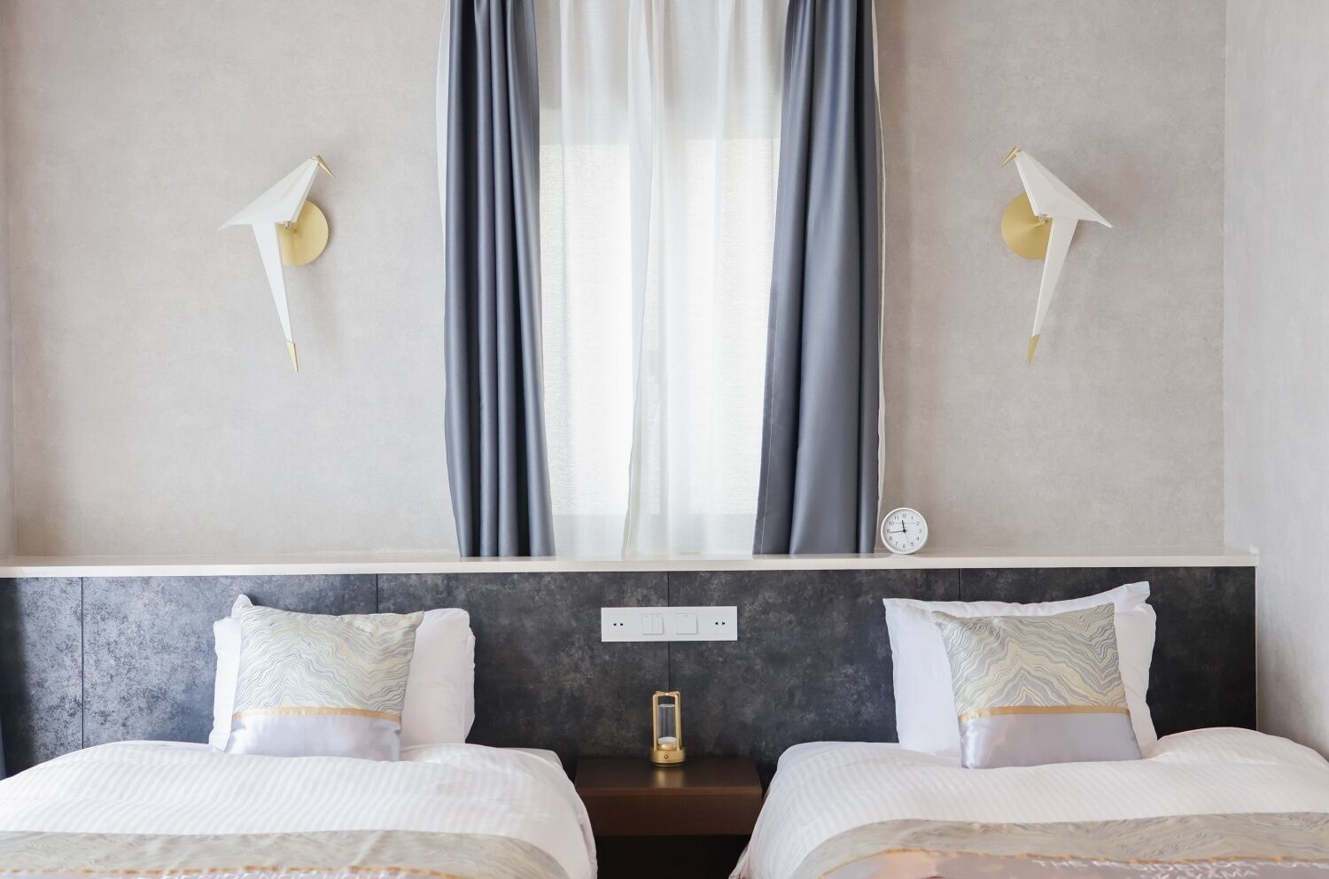 Ein Hotelzimmer mit zwei Betten, einem großen Fenster mit Vorhängen, einer Wanduhr und zwei Wandleuchten.