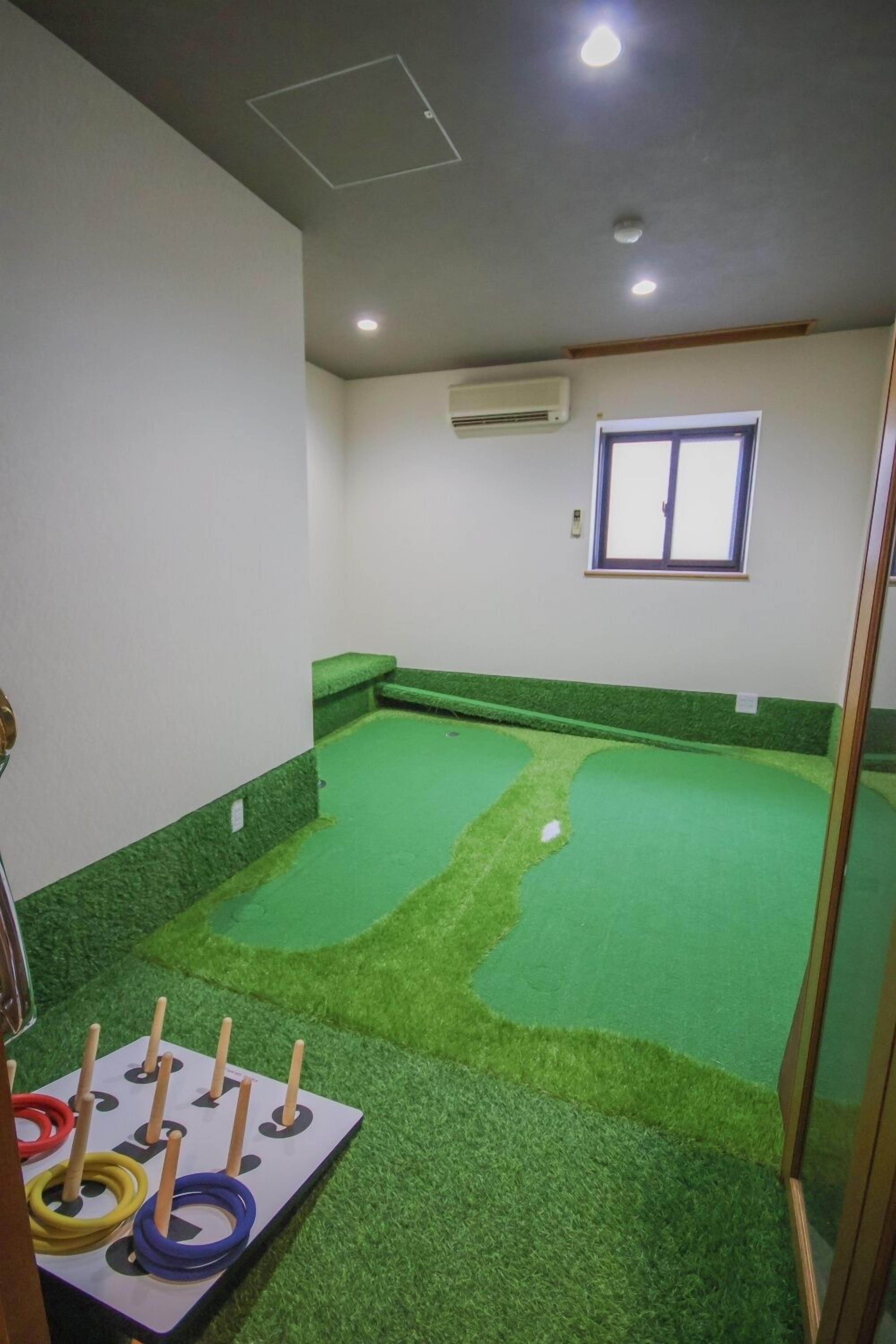 Una habitación con una zona de césped artificial verde, una mesita con anillos de colores y una ventana con persianas.