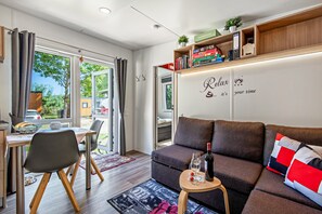 Un salon moderne comprenant un canapé, une petite table avec une bouteille et un verre, un coin repas avec des chaises et une table, ainsi qu’une bibliothèque remplie de livres et d’objets décoratifs.