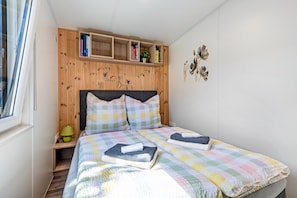 Ein Schlafzimmer mit einem hölzernen Kopfteil, einem Bett mit farbenfroher Decke, einem Nachttisch mit Lampe und einem Regal mit Büchern und Dekorationsgegenständen.