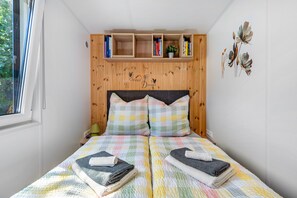 Un lit avec une couverture à carreaux, deux oreillers et des serviettes pliées. Il y a une tête de lit en bois avec des étagères au-dessus du lit.